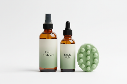 Amber spray bottle and amber dropper bottle with black caps, both with vertical cream-to-sage gradient labels and black serif text that say ‘Hair Freshener’ and ‘Liquid Gold.’ Light green massage bar with 13 bumps beside them. Clean studio background